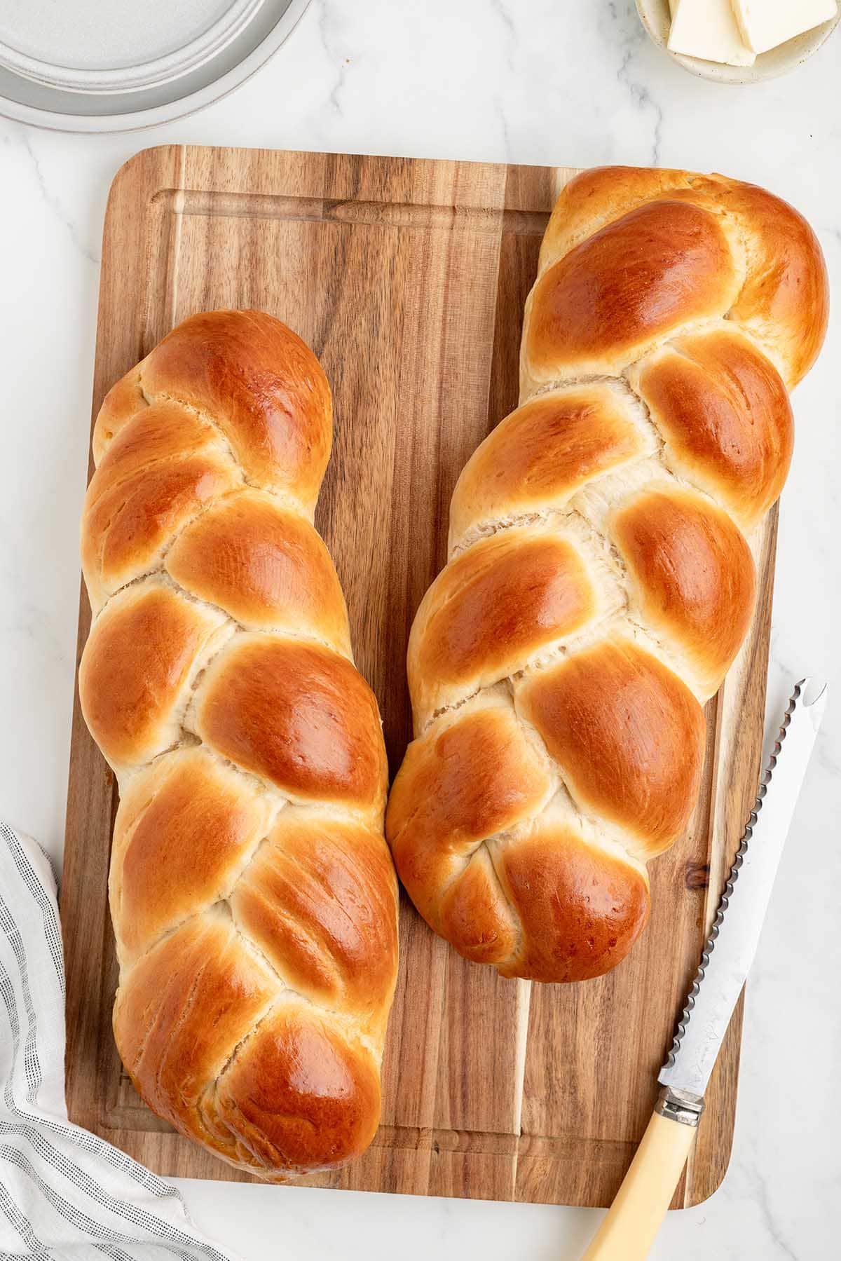 Two loaves of challah bread on a wooden cutting board with a serrated knife.