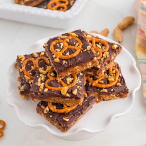 a stack of chocolate peanut butter pretzel bars topped with pretzels and crushed peanuts served on a white plate.