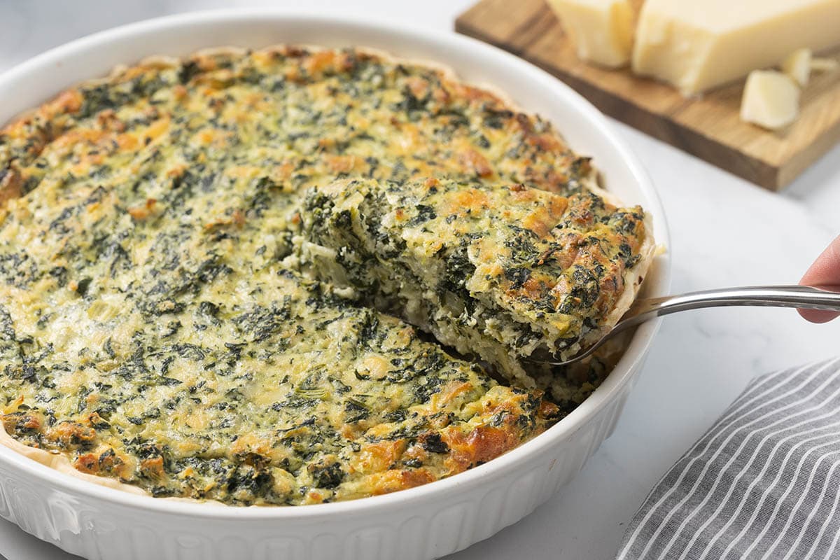 Spinach pie in a white baking dish with a slice being lifted out, showing a cheesy spinach filling.