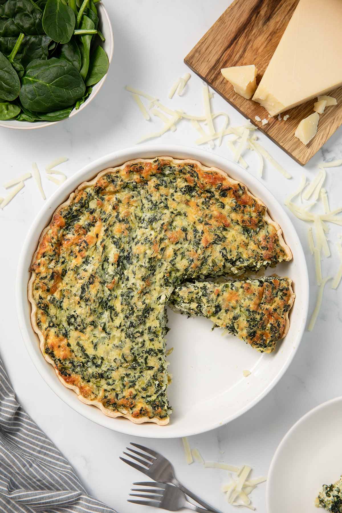 Spinach pie in a white dish with a slice removed, surrounded by fresh spinach and parmesan cheese.