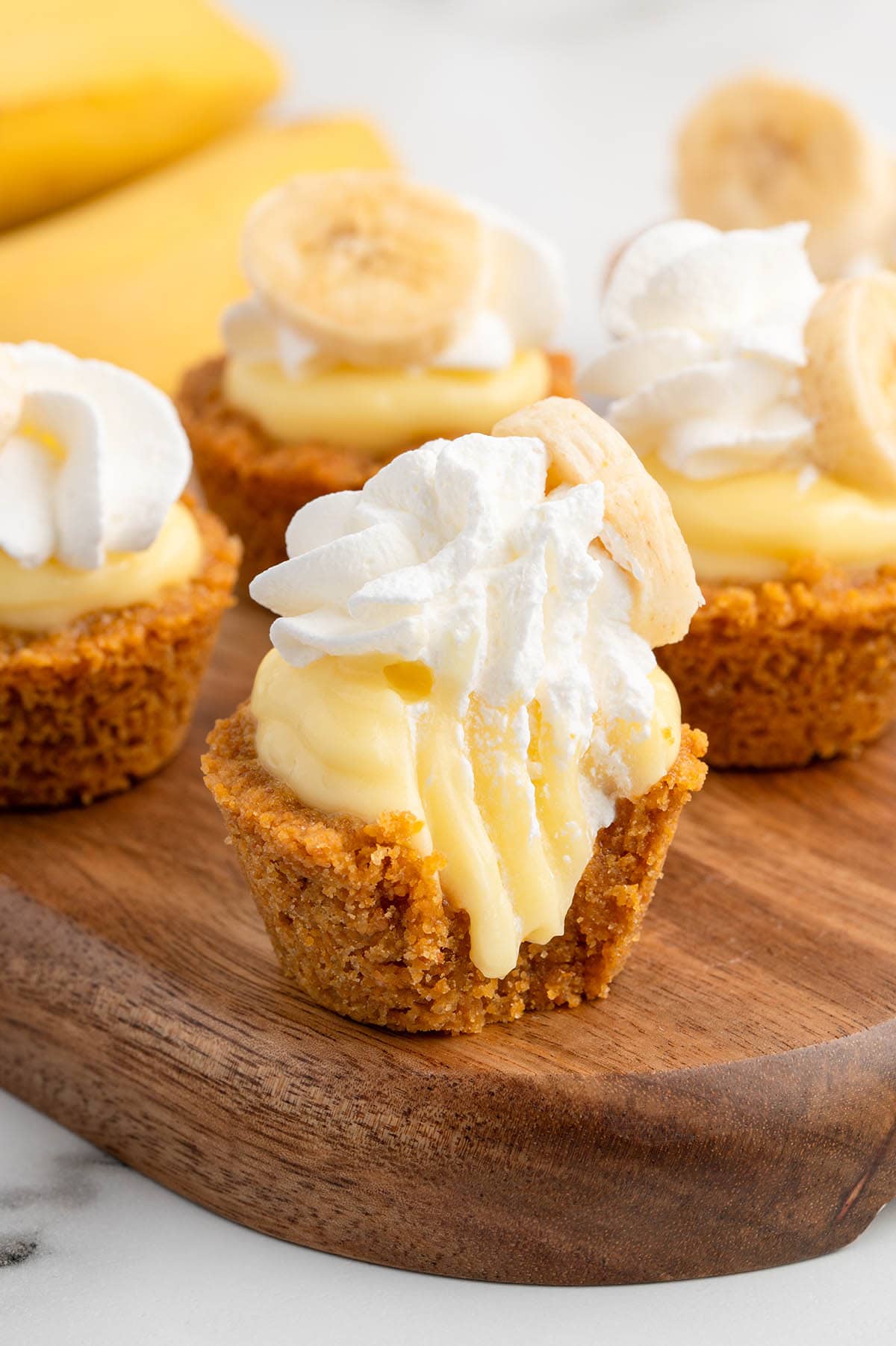 Four mini banana cream pies on a wooden board, topped with whipped cream and banana slices.
