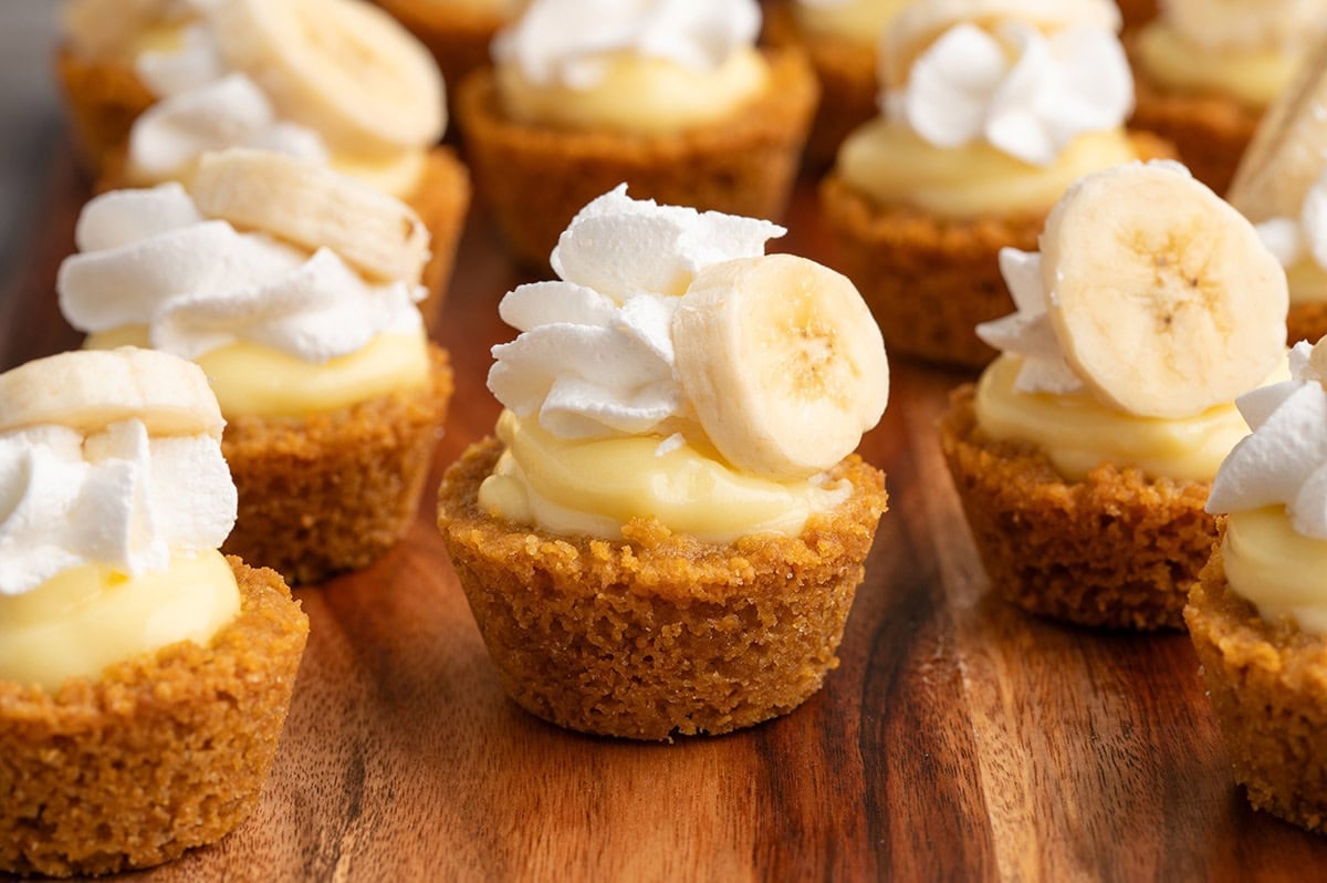 Mini banana cream pies with graham cracker crusts, custard filling, whipped cream, and banana slices on a wooden board.