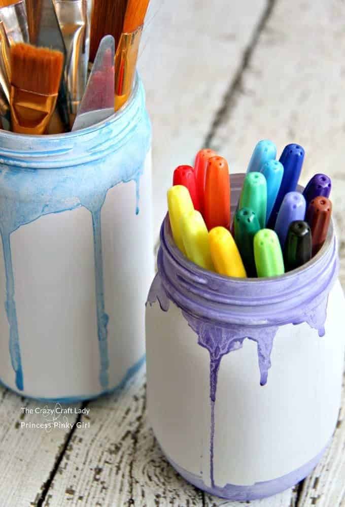 A mason jar painted with white paint and dripping with purple and blue paint. Filled with markers.