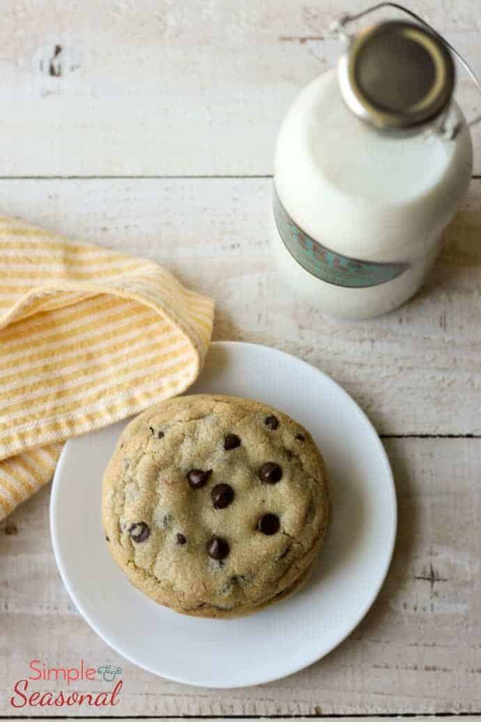 A chocolate chip cookie sitting on a white plate with yellow and white linens around it and a glass of milk