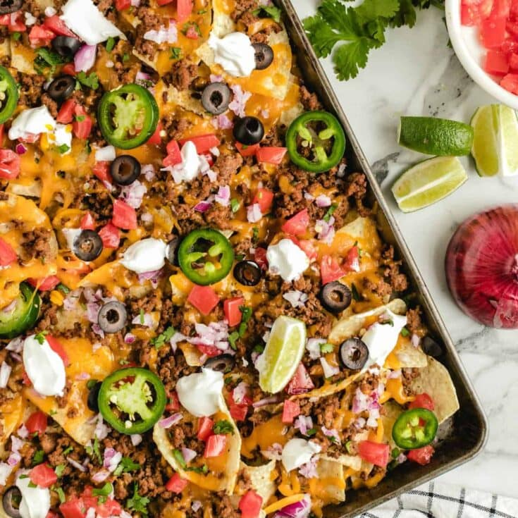 Sheet pan nachos topped with ground beef, melted cheese, tomatoes, onions, olives, jalapenos, sour cream, and cilantro.