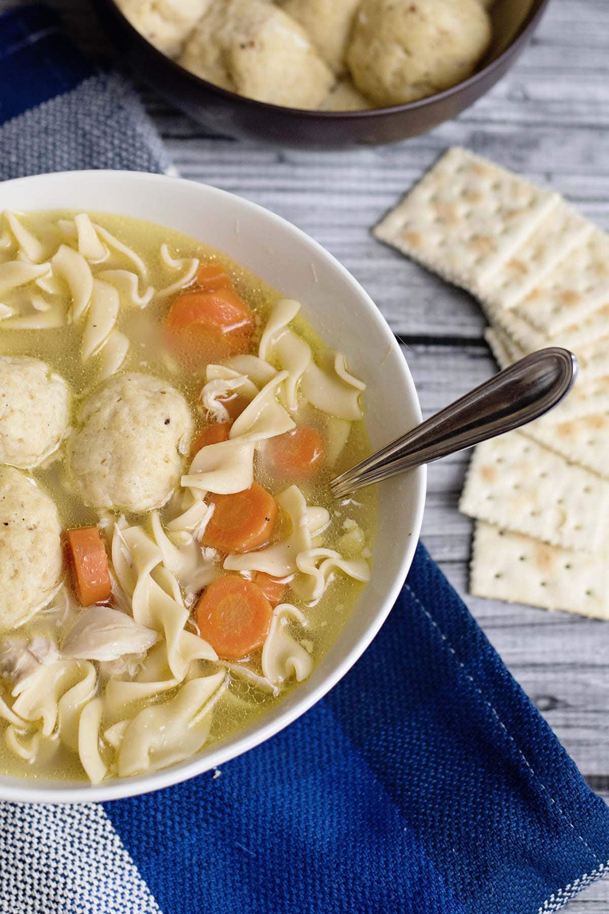 Bowl of chicken soup with egg noodles, carrots, and matzo balls, served with crackers on a blue napkin for the Chicken Soup Recipe.