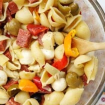 A glass bowl filled with pasta salami peppers olives and cheese