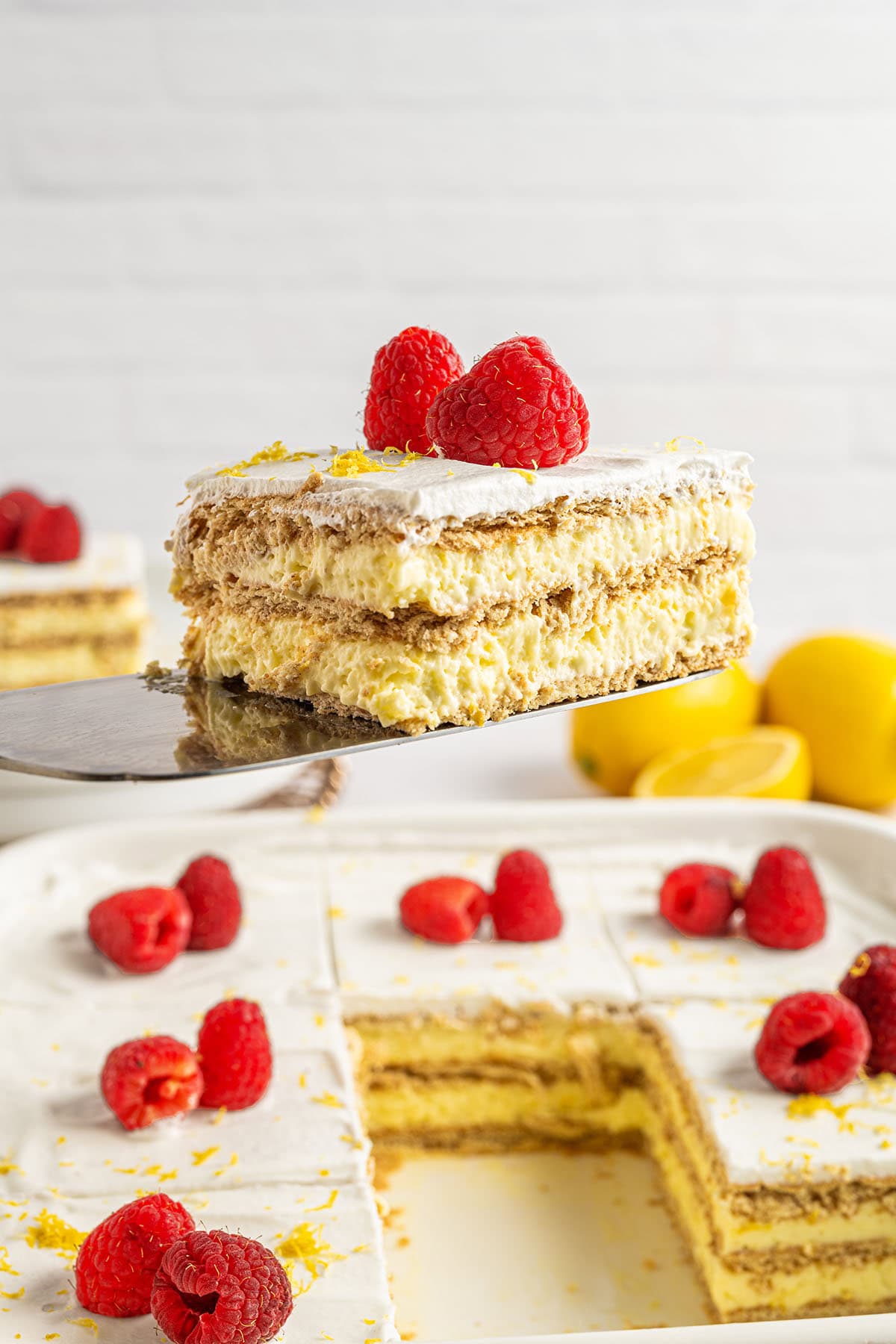 A slice of lemon icebox cake with whipped topping, graham cracker layers, and fresh raspberries, lifted from a baking dish.