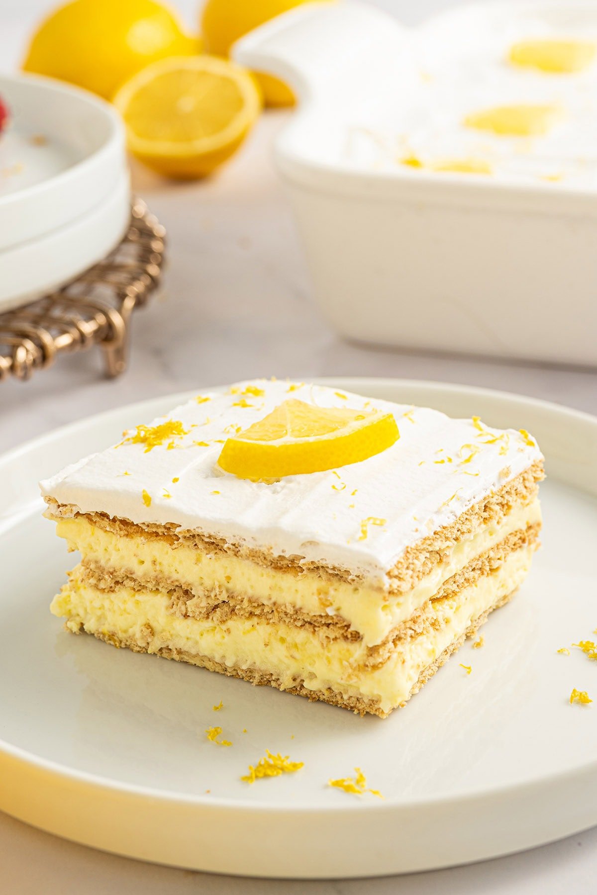 Slice of lemon icebox cake topped with a lemon wedge and lemon zest, layered with graham crackers and creamy lemon filling on a white plate.