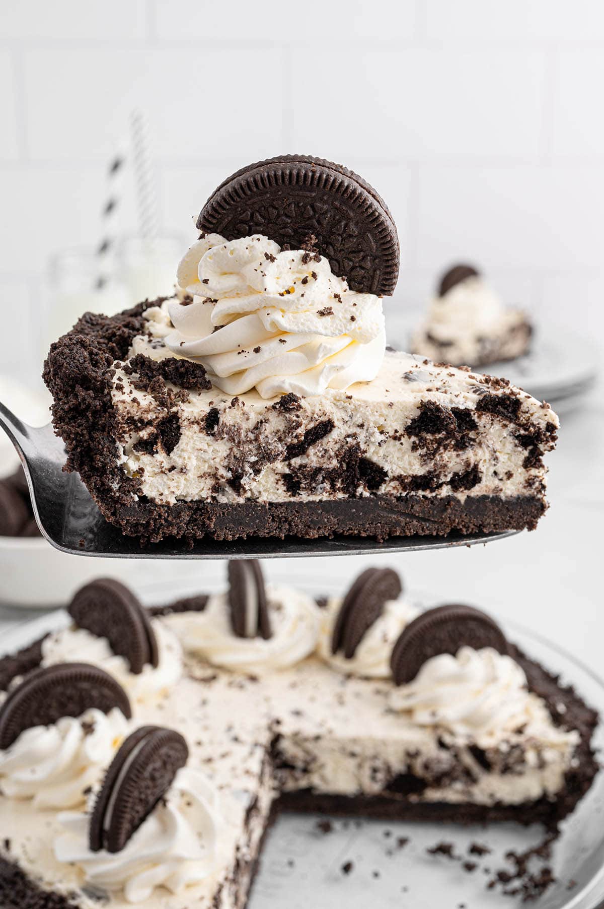 A slice of No Bake Oreo Pie being lifted on a server, showcasing the creamy Oreo filling, whipped topping, and cookie garnish, with the rest of the pie in the background.