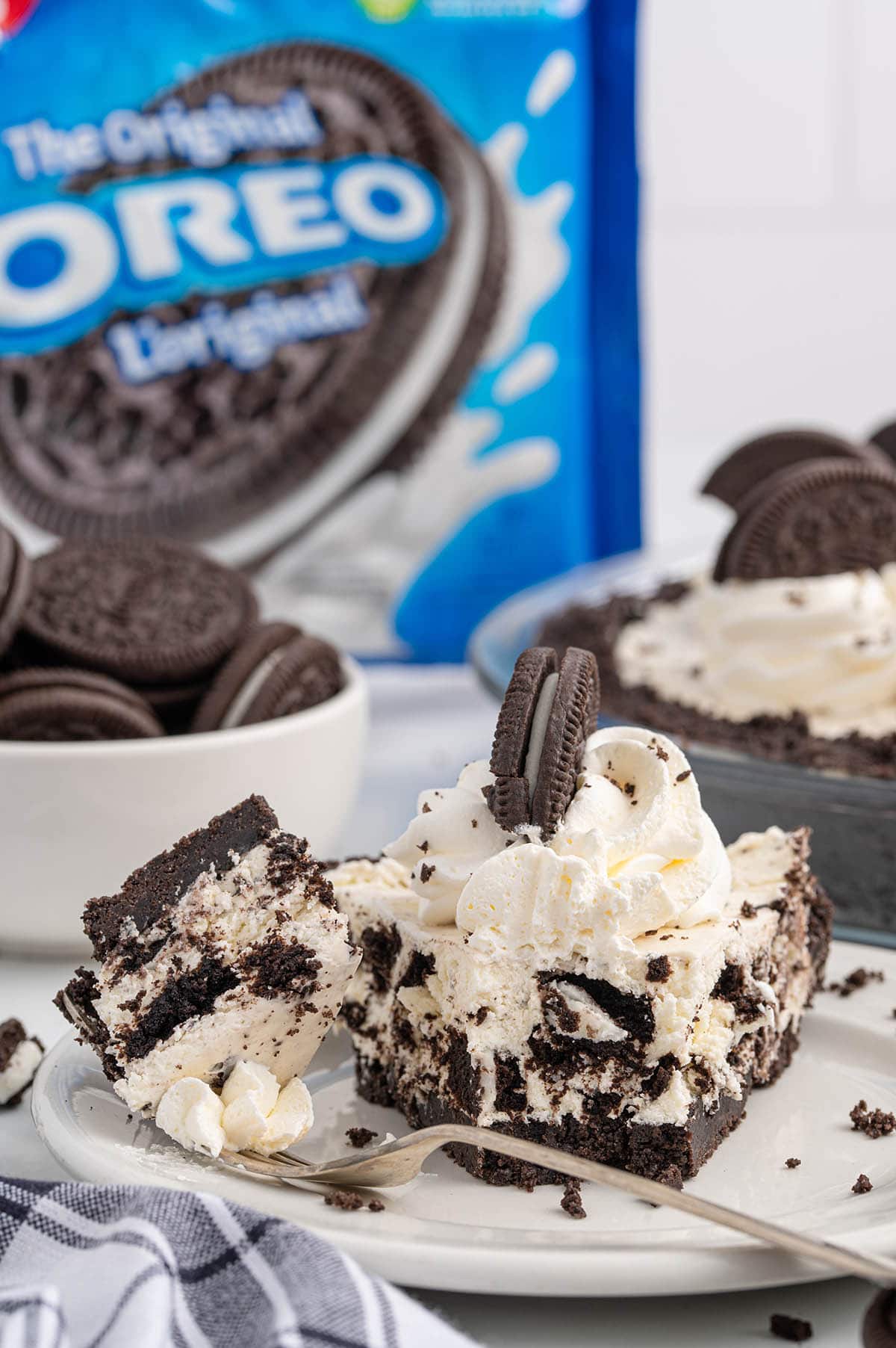 Slice of No Bake Oreo Pie on a plate with a fork, topped with whipped cream and an Oreo cookie, with an Oreo package in the background.