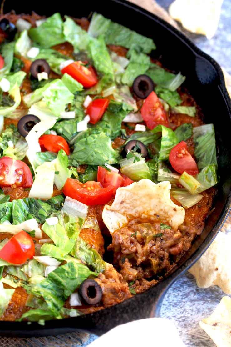 A close-up image of taco dip with chips tomatoes lettuce in a skillet