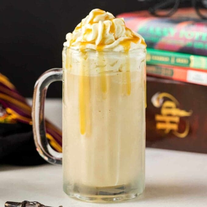 A glass filled with Butterbeer with whipped cream and caramel sauce dripping down the side of the glass. Harry Potter books in the background. 