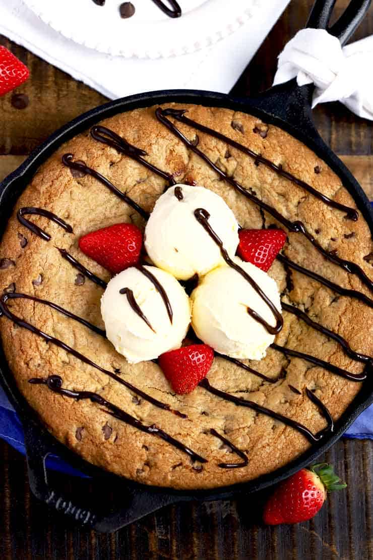 Chocolate chip cookie skillet with three scoops of vanilla ice cream and strawberries on top drizzled with hot fudge