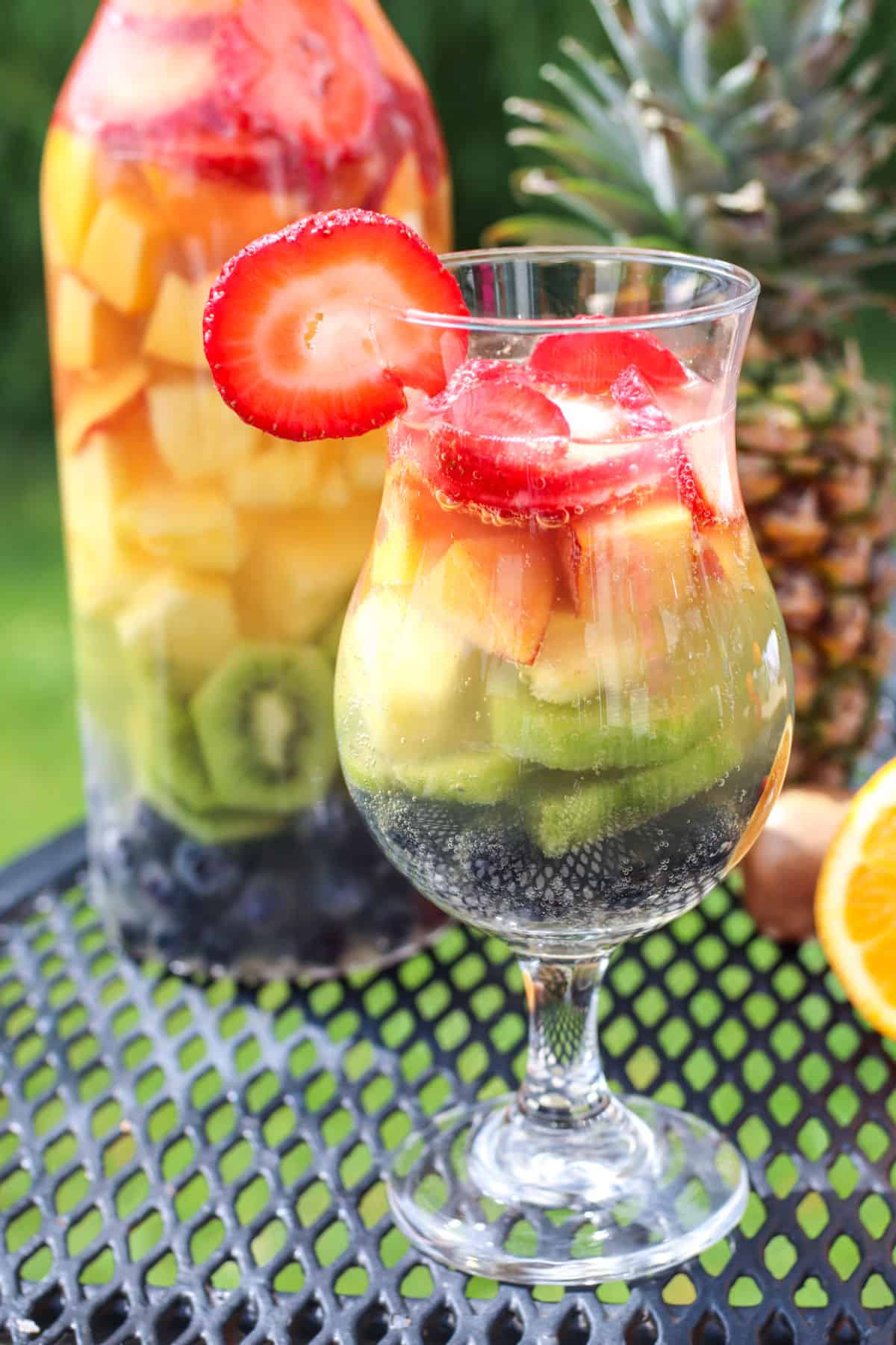 A glass of sangria with different fruits with a strawberry on the side outside on a wire table