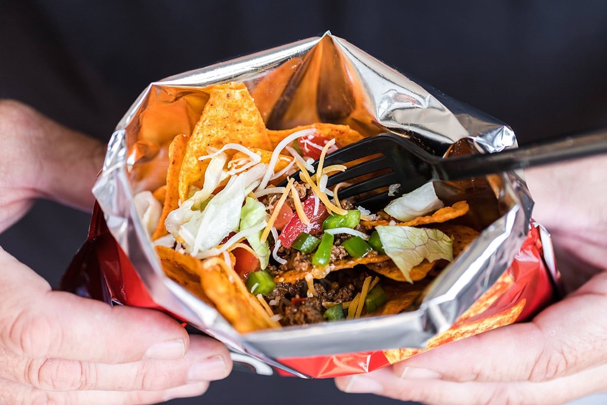 Close-up of hands holding a bag of Nacho Cheese Doritos filled with taco ingredients, including lettuce, cheese, tomatoes, and ground beef, with a black fork inside.