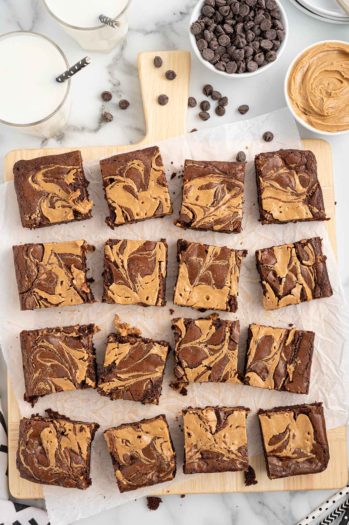 peanut butter brownies with a swirl design, cut into squares on parchment paper, surrounded by glasses of milk, chocolate chips, and peanut butter.