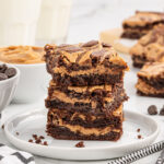 A stack of peanut butter brownies on a plate, with bowls of chocolate chips and peanut butter in the background.