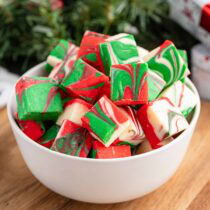 Red, green, and white swirled Christmas fudge squares piled in a white bowl with evergreen branches in the background.