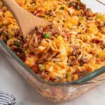 Wooden spoon scooping cheesy hamburger noodle casserole from a glass baking dish.