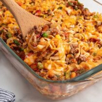 Wooden spoon scooping cheesy hamburger noodle casserole from a glass baking dish.