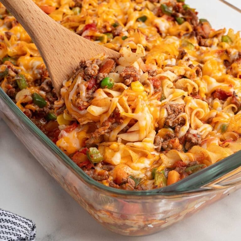 Wooden spoon scooping cheesy hamburger noodle casserole from a glass baking dish.