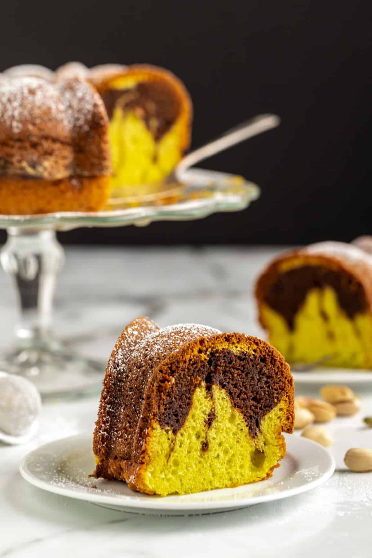 a slice of chocolate pistachio cake on a white plate with the full cake in the background