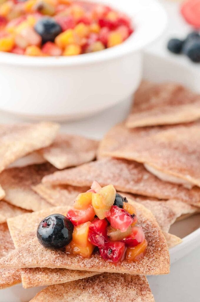 fruit salsa on top of a cinnamon chip with more chips and a white bowl of salsa in the background