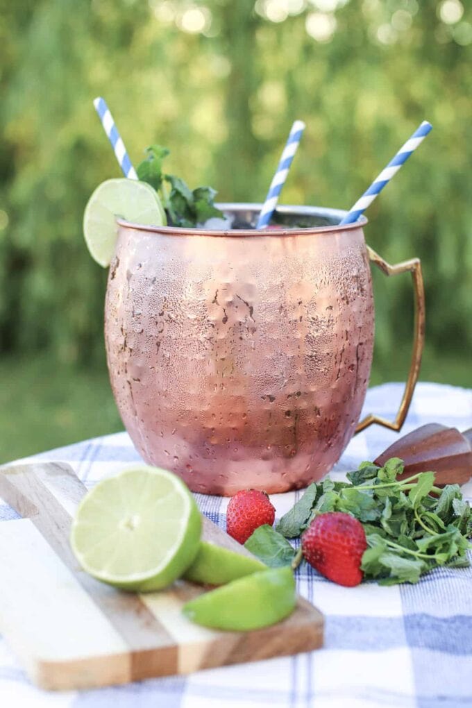 Moscow mule in a copper mug with striped straws and limes and strawberries on the table with mint leaves