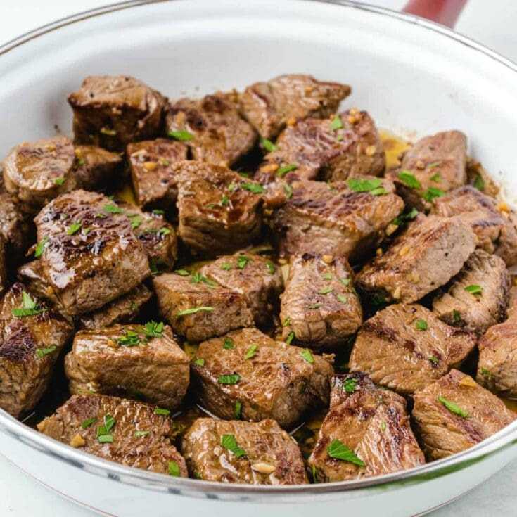 Garlic butter steak bites sizzle in a white pan topped with parsley.