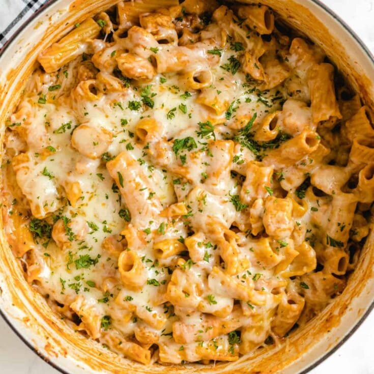 Close-up of one pot chicken parmesan pasta in a white pot, with creamy tomato sauce, melted cheese, and fresh parsley on top.