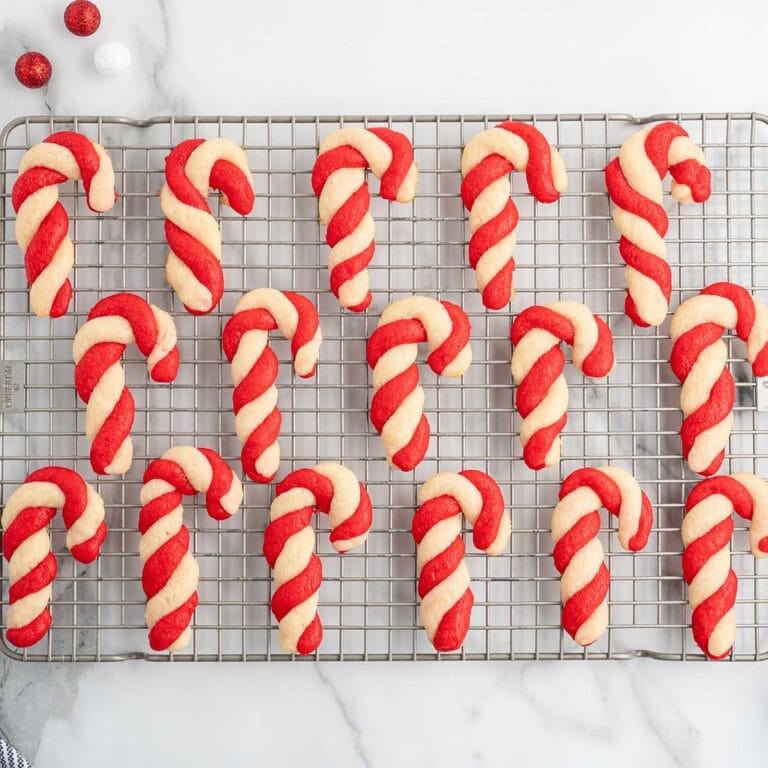 Rows of red and white candy cane-shaped cookies on a wire cooling rack, with ornaments nearby.