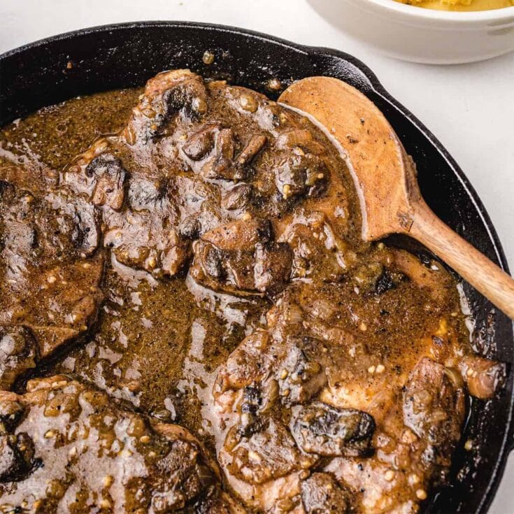 Smothered pork chops in mushroom gravy in a cast iron skillet with a wooden spoon.