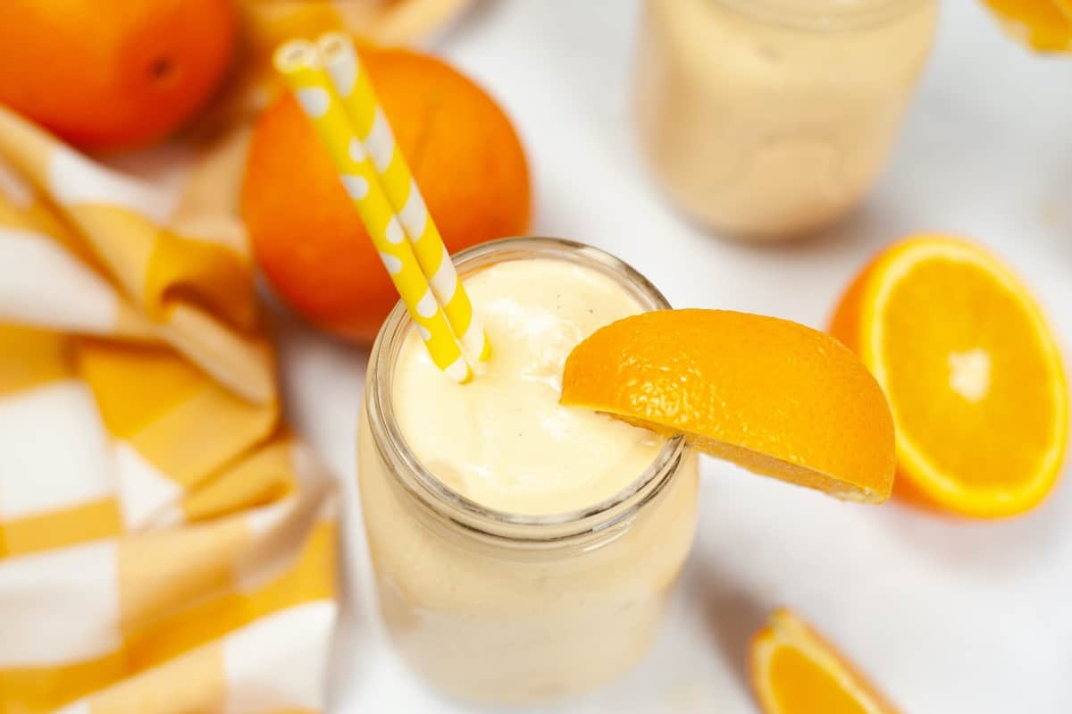Top view of a creamy orange smoothie in a mason jar with an orange slice garnish, surrounded by fresh oranges and a yellow striped cloth.
