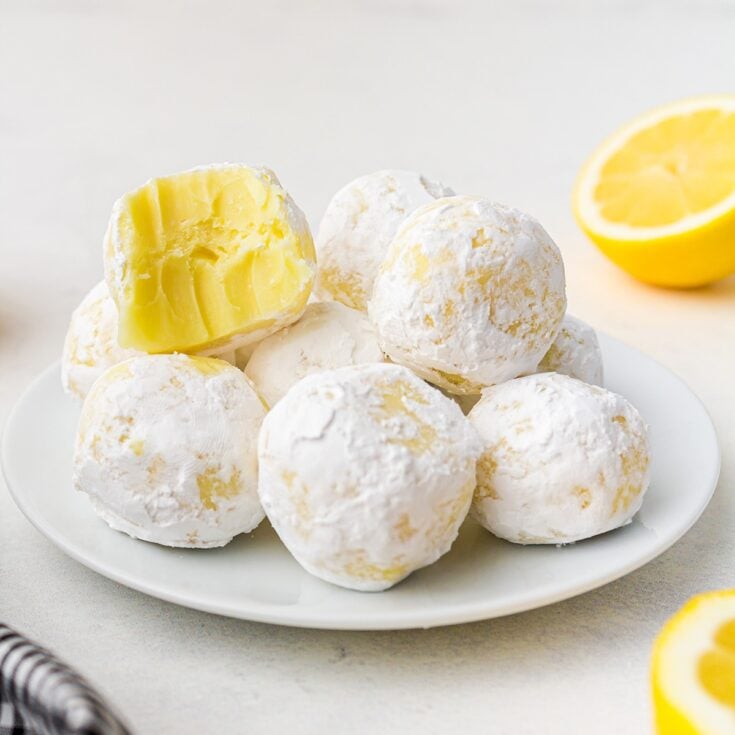 White chocolate lemon truffles coated in powdered sugar, stacked on a white plate with one truffle showing a creamy lemon filling.