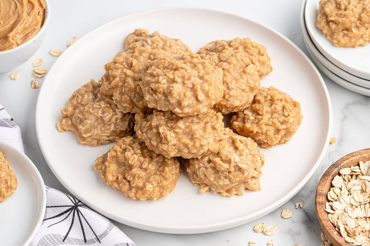 A plate stacked with peanut butter no-bake cookies made with oats, surrounded by bowls of oats and peanut butter on a white surface.