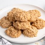 Plate of peanut butter no-bake cookies made with oats, stacked neatly on a white dish with a few scattered oats nearby.