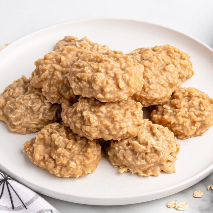 Plate of peanut butter no-bake cookies made with oats, stacked neatly on a white dish with a few scattered oats nearby.
