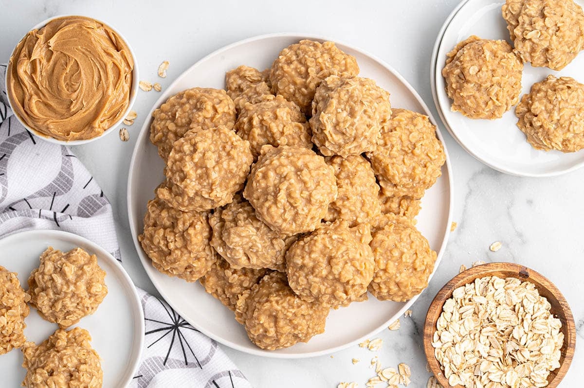 Overhead view of a white plate stacked with peanut butter no-bake cookies, surrounded by bowls of oats and peanut butter, with additional cookies on side plates.
