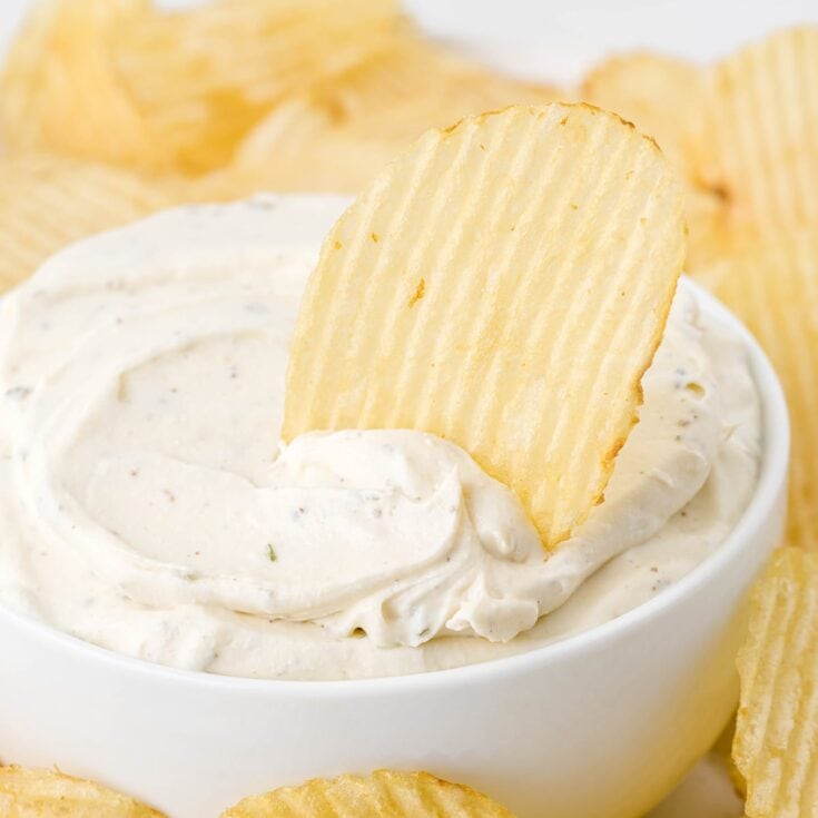 Ridged potato chip dipped into a creamy chip dip in a white bowl, surrounded by more chips.