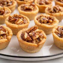 Mini pecan pies with flaky crusts topped with pecan halves, arranged on a white platter.