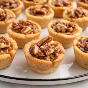 Mini pecan pies with flaky crusts topped with pecan halves, arranged on a white platter.