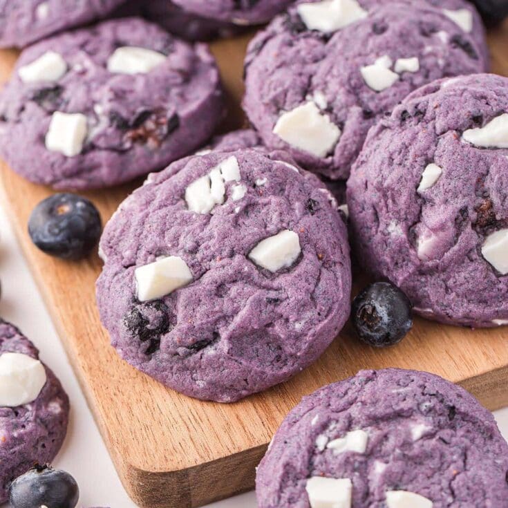 Vibrant purple blueberry cookies with chunks of white chocolate, arranged on a wooden board with fresh blueberries scattered around.
