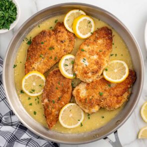 Lemon chicken in a skillet with sauce, parsley, and lemon slices.