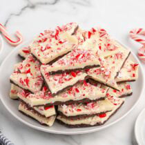 Peppermint bark pieces topped with crushed candy cane, piled on a white plate with candy canes nearby.
