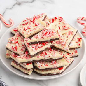 Peppermint bark pieces topped with crushed candy cane, piled on a white plate with candy canes nearby.