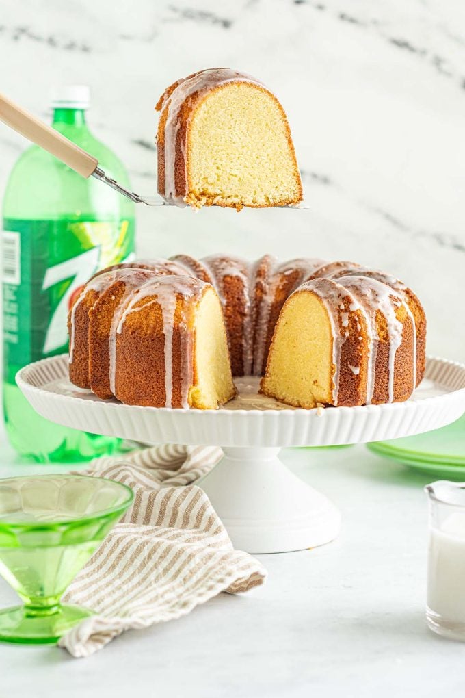 A slice of 7Up Pound Cake being lifted from a glazed bundt cake on a white cake stand, with a bottle of 7Up in the background.