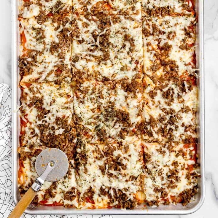 Overhead view of a sheet pan pizza cut into squares, topped with melted cheese and ground beef, next to a pizza cutter.