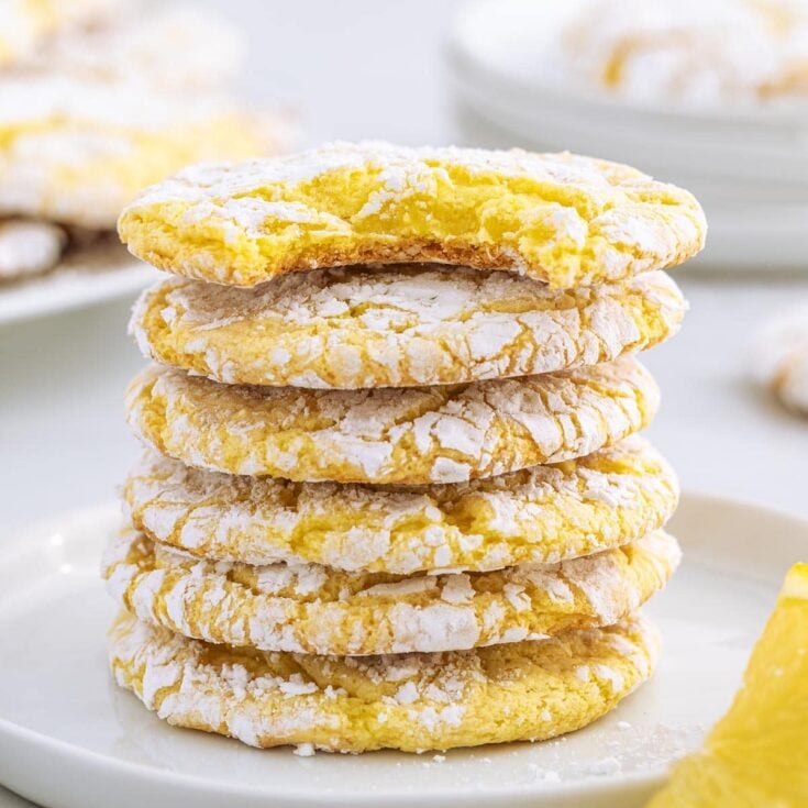A stack of five lemon cool whip cookies topped with powdered sugar sits on a white plate next to a fresh lemon slice.