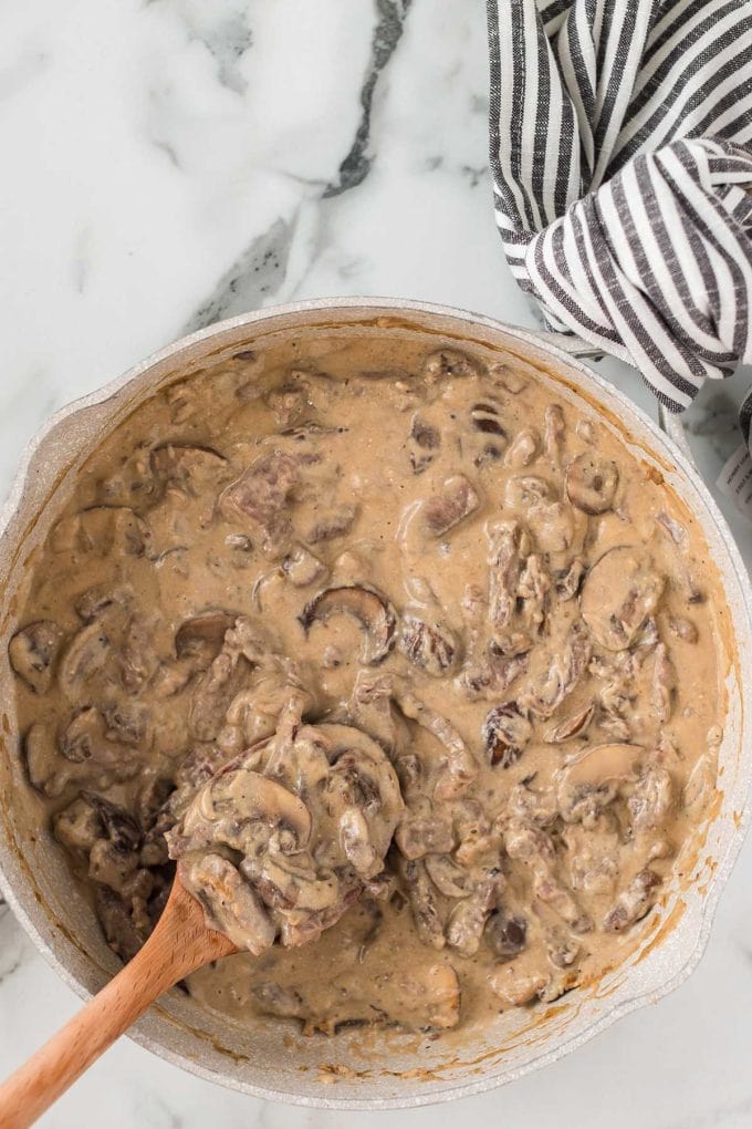 Creamy beef stroganoff fills a white pot. Mushrooms and beef chunks swim in thick sauce. A wooden spoon stirs it on marble.